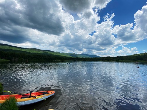 A serene lake surrounded by green hills and fluffy clouds, with a kayak resting on the shore.