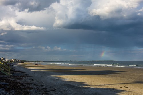 A beach scene with cloudy skies, a faint rainbow, and people walking along the shore. Shadows stretch across the sand.