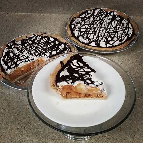 A slice of apple pie on a plate, with two whole pies in the background, topped with chocolate drizzle and whipped cream.