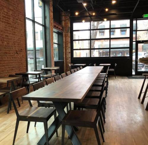 A spacious restaurant interior featuring a long wooden table and chairs, with large windows and exposed brick walls.