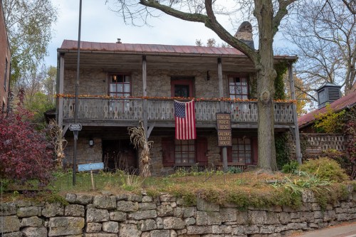 Historic stone house with a wooden balcony, American flag, and autumn decorations, surrounded by greenery and stone walls.