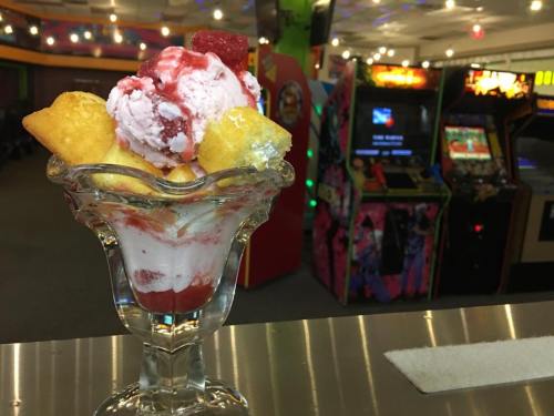 A glass dessert dish filled with strawberry ice cream, fruit, and cake pieces, with an arcade in the background.