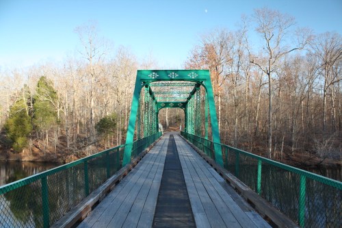 A green metal bridge spans a calm river, surrounded by bare trees and a clear blue sky.