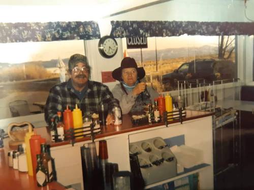 Two people sit at a diner counter, surrounded by condiments, with a scenic view outside the window.