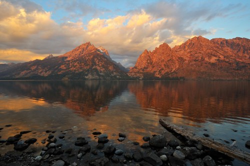 Sunset over a tranquil lake, reflecting rugged mountains and colorful clouds, with a rocky shoreline in the foreground.
