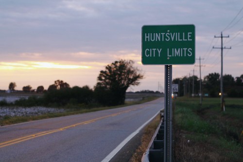 Sign marking the city limits of Huntsville, with a rural road and cloudy sky in the background.