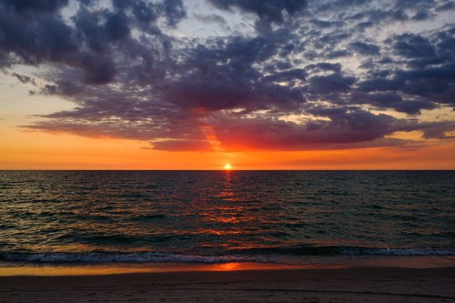 Sunset over the ocean, with vibrant orange and purple hues reflecting on the water and clouds scattered in the sky.