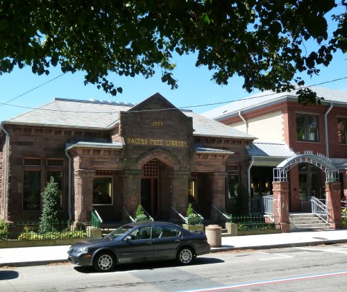 Historic Rogers Free Library building with stone facade, surrounded by trees and a parked car in front.