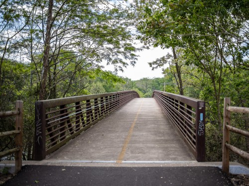 A pedestrian bridge surrounded by trees, leading into a green landscape under a cloudy sky.