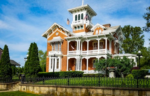 A grand, historic brick mansion with a tower, white trim, and lush greenery, set against a blue sky.