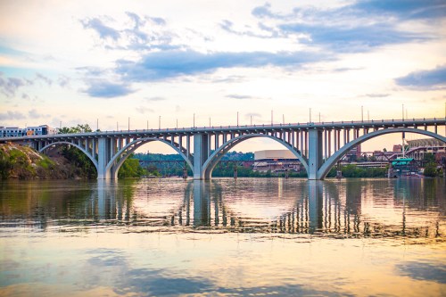 A bridge spans a river at sunset, reflecting its arches in the calm water below. Soft clouds fill the sky.