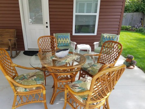 A patio set with a glass table and six wicker chairs, surrounded by a green lawn and a wooden wall.