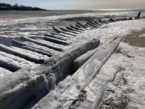 Frozen wooden structures along a snowy beach, with ice formations glistening in the sunlight.
