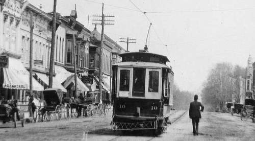 Historic street scene featuring a trolley car, horse-drawn carriages, and early 20th-century buildings.