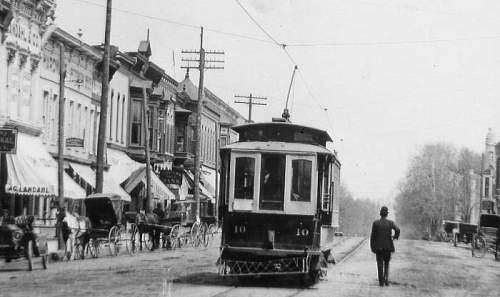 Historic street scene featuring a trolley car, horse-drawn carriages, and early 20th-century buildings.