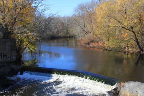 A serene river scene with a small waterfall, surrounded by autumn trees and calm waters reflecting the landscape.