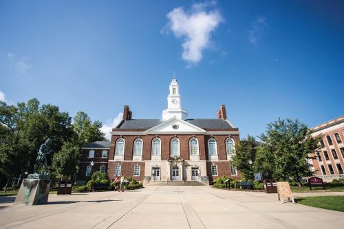 A brick building with a clock tower, surrounded by trees and a clear blue sky, in a campus setting.