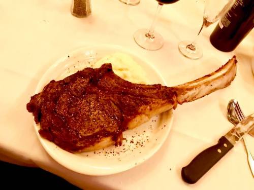 A large, juicy tomahawk steak on a white plate, served with mashed potatoes and accompanied by wine glasses.