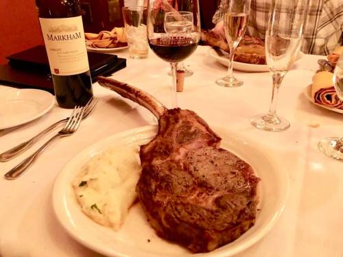 A large ribeye steak on a plate with mashed potatoes, accompanied by a bottle of red wine and glasses on a table.