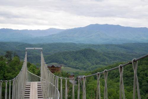 A suspension bridge stretches over lush green mountains under a cloudy sky, leading to distant peaks.