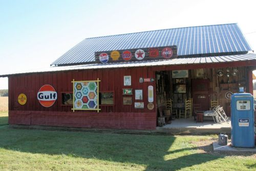 A rustic red building with vintage Gulf signs and a gas pump, surrounded by green grass and fields.