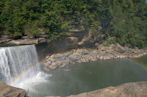 A waterfall cascades into a river, creating mist and a rainbow against a backdrop of lush green trees and rocky cliffs.