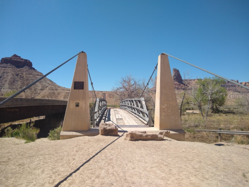 A suspension bridge spans a dry riverbed, surrounded by rocky formations and sparse vegetation under a clear blue sky.