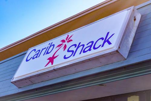 Sign for "Carib Shack" featuring palm tree graphics, mounted on a building with a clear blue sky above.