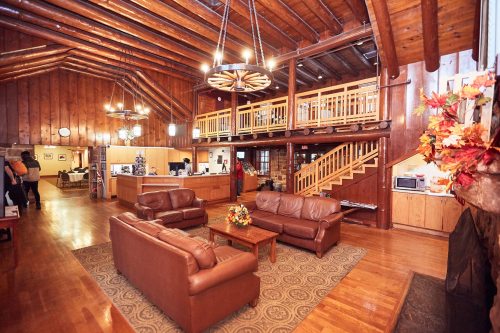 Cozy lodge interior with wooden beams, leather sofas, a coffee table, and a staircase leading to a balcony.