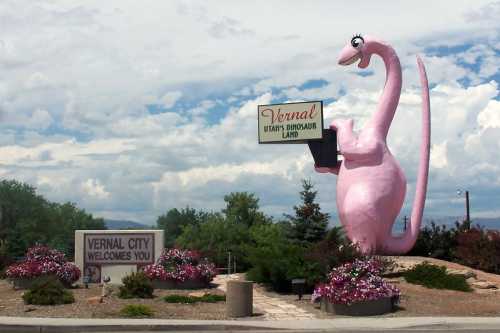 A large pink dinosaur statue holds a sign for Vernal, Utah, surrounded by flowers and welcoming signs.