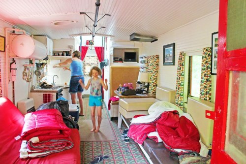 A cozy interior of a small cabin with a woman cooking and a child playing, surrounded by colorful decor and bedding.
