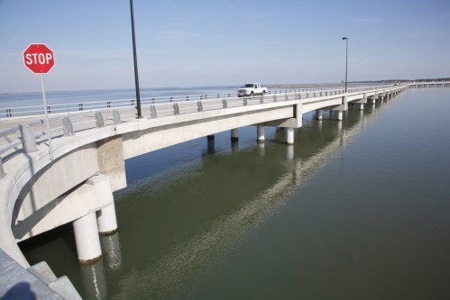 A concrete bridge extends over calm water, with a stop sign visible and a vehicle driving on the bridge.