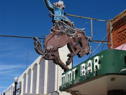 Neon sign of a cowboy riding a bucking horse above the Mint Bar, with a clear blue sky in the background.