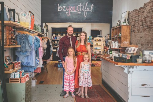 A family of four stands in a boutique, smiling in front of a "beYOUtiful" sign, surrounded by clothing and decor.