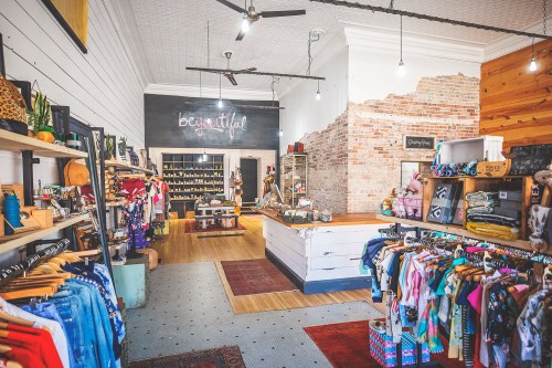 A stylish boutique interior featuring clothing racks, wooden shelves, and a rustic brick wall. Bright and inviting atmosphere.