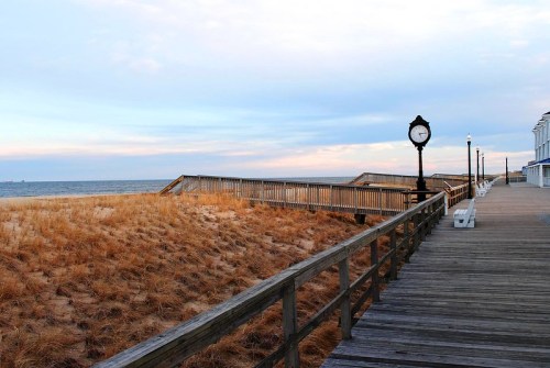 A wooden boardwalk along a beach, with a clock and grass-covered dunes under a cloudy sky.