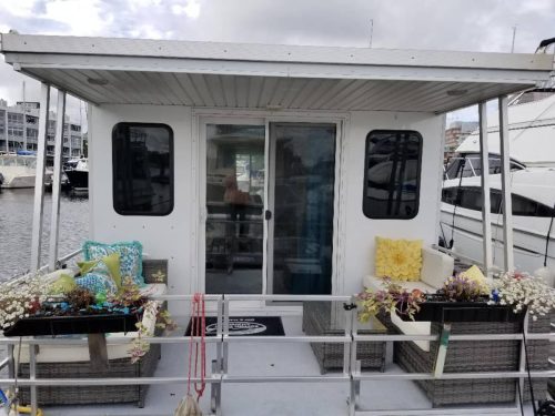 A cozy houseboat with a covered deck, featuring potted plants and comfortable seating, docked in a marina.