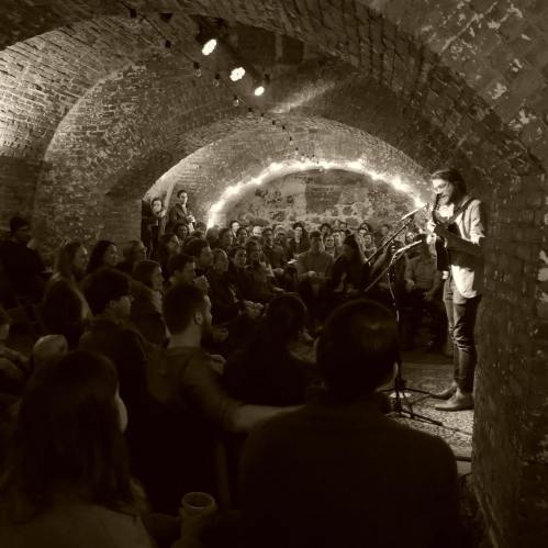 A performer plays guitar in a dimly lit, arched brick venue, while an engaged audience listens intently.