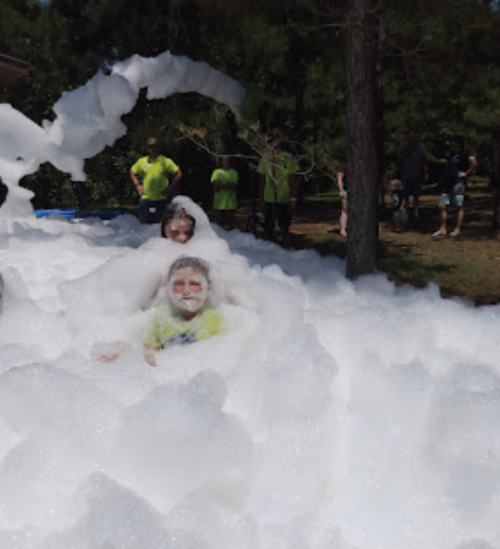 Two children play in a large pile of foam, with adults in bright shirts visible in the background among trees.