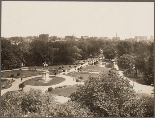 A historic park scene featuring a statue, lush greenery, and a winding path, with city buildings in the background.