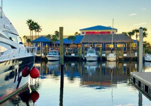 A marina at sunset with boats docked and a restaurant in the background, reflecting in the calm water.