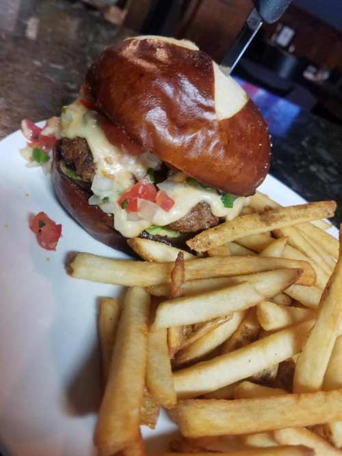 A pretzel bun burger topped with cheese, tomatoes, and green onions, served with a side of golden fries.