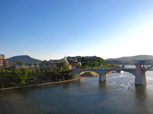 A scenic view of a river with a bridge, surrounded by greenery and buildings under a clear blue sky.