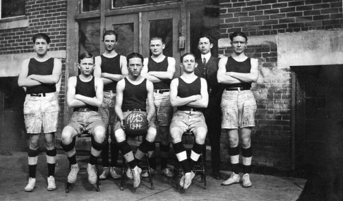 A vintage basketball team poses for a photo, wearing uniforms and holding a basketball, with a coach standing behind them.