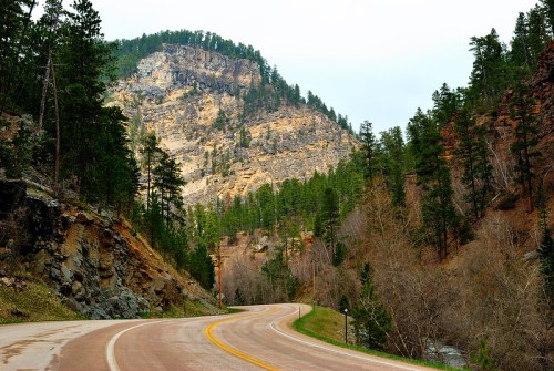 Winding road through a forested landscape with rocky cliffs and mountains in the background.