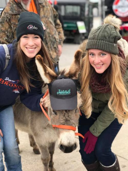 Two women smile beside a donkey wearing a cap, with a festive outdoor setting in the background.