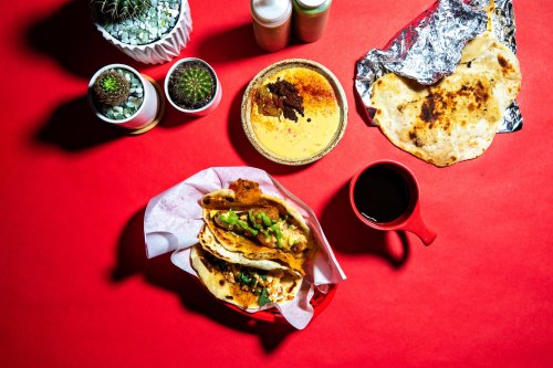 A vibrant red table with tacos, a bowl of sauce, a cup of coffee, and small potted cacti.
