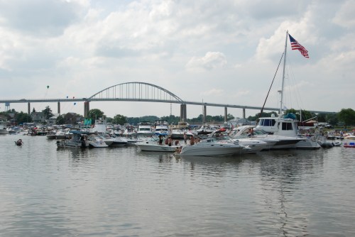 A busy marina with various boats on the water, featuring a bridge and an American flag in the background.