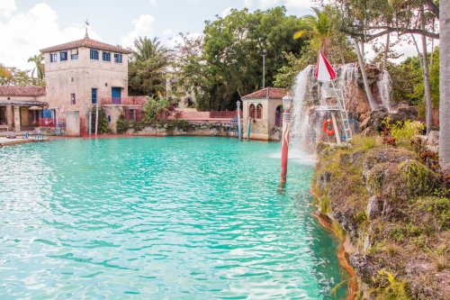 A serene turquoise pool with a waterfall, surrounded by lush greenery and a rustic building in the background.