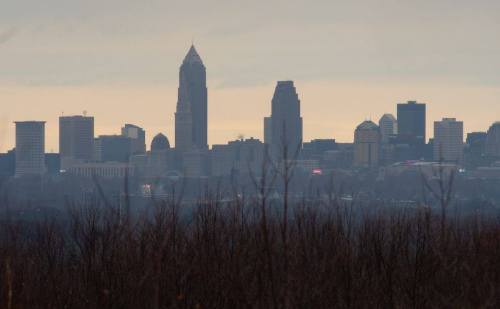 Silhouette of a city skyline with tall buildings against a cloudy sky, viewed through bare tree branches.